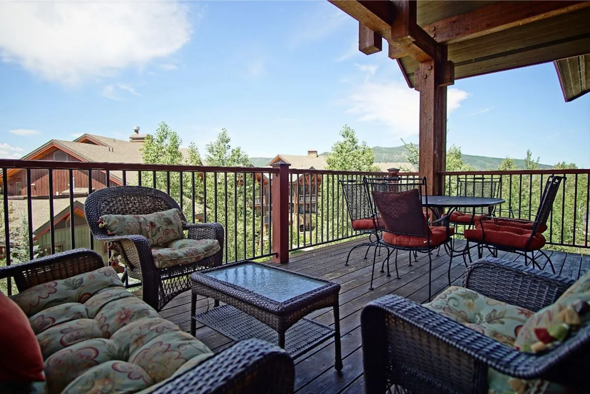 A spacious outdoor deck furnished with comfortable seating, a wooden railing, and a glass coffee table. The scene is set against a backdrop of lush trees and distant mountains, suggesting a tranquil retreat perfect for relaxation or gathering with friends and family. The golden glow hints at sunset or early morning, with soft light filtering through the trees.