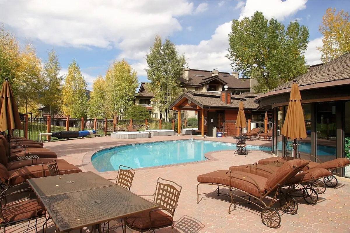 This lively image features a sunlit swimming pool surrounded by lounge chairs, patio umbrellas, and trees, with guests enjoying sunbathing, swimming, and socializing in a cheerful outdoor setting.