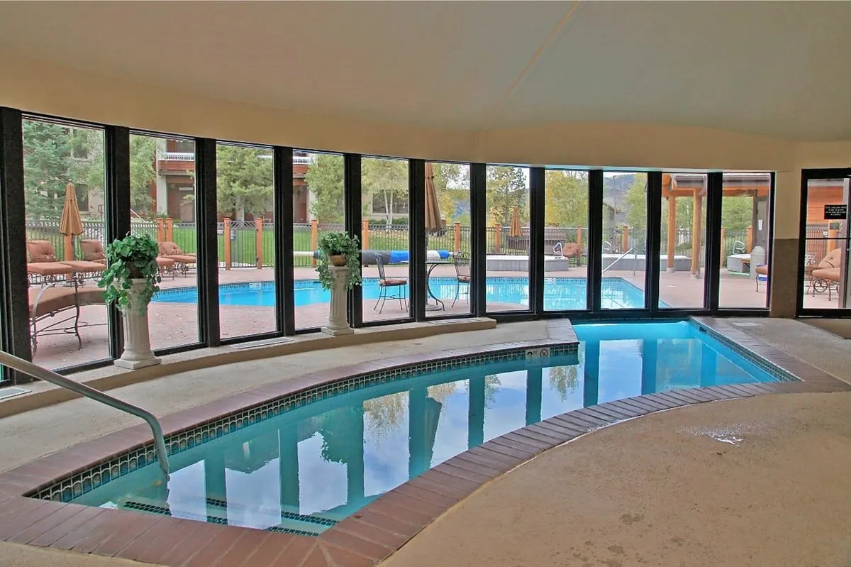 An inviting interior space featuring a large indoor hot tub, surrounded by ample windows that reveal the outdoor pool deck. Soft lighting and decorative plants create a tranquil, spa-like atmosphere perfect for relaxation and wellness. The image provides a clear view of both indoor and outdoor amenities.