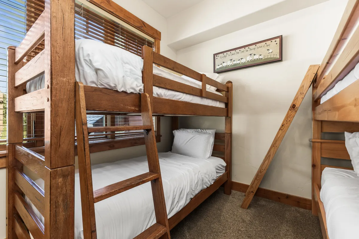 A playful bunk room featuring sturdy wooden bunk beds, a wooden ladder, and a window with blinds. The room is decorated with cheerful wall art, creating a lively and inviting atmosphere perfect for children. The space encourages both fun and relaxation, with ample room for games or bedtime stories.