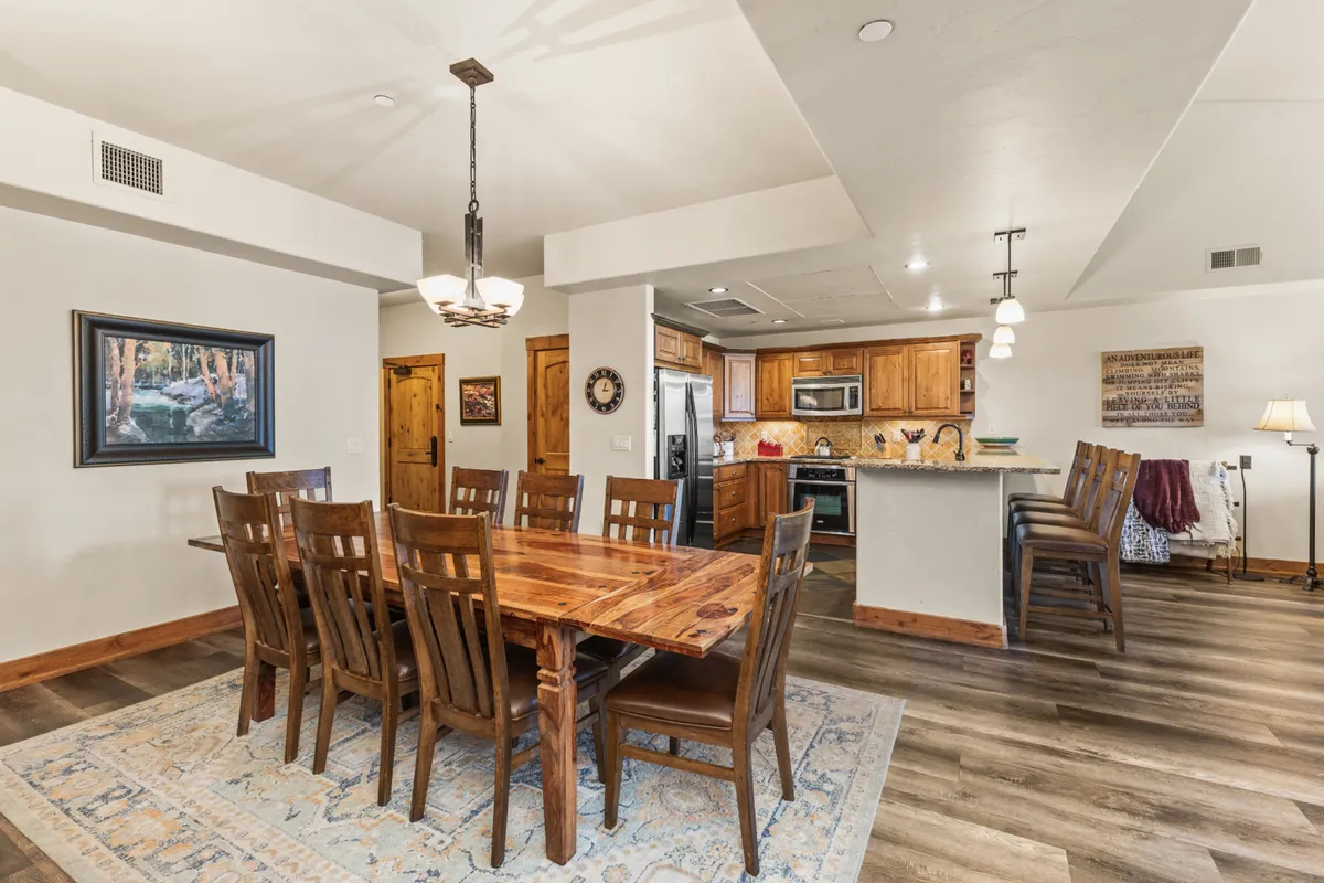 The focus is on a spacious wooden dining table with comfortable chairs, set beneath a stylish chandelier. The kitchen area with matching wooden cabinets is visible in the background, seamlessly integrating cooking and dining spaces. The mood is warm and welcoming, perfect for family meals or entertaining friends.