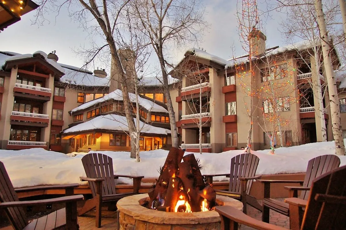 This image presents an inviting outdoor fire pit surrounded by chairs, set beside the building's facade and under trees strung with lights. The atmosphere is especially magical, suggesting a gathering space where people share stories, enjoy drinks, and relax together after a day outdoors. Visible snow hints at winter festivities, making it a perfect representation of Après Ski parties.