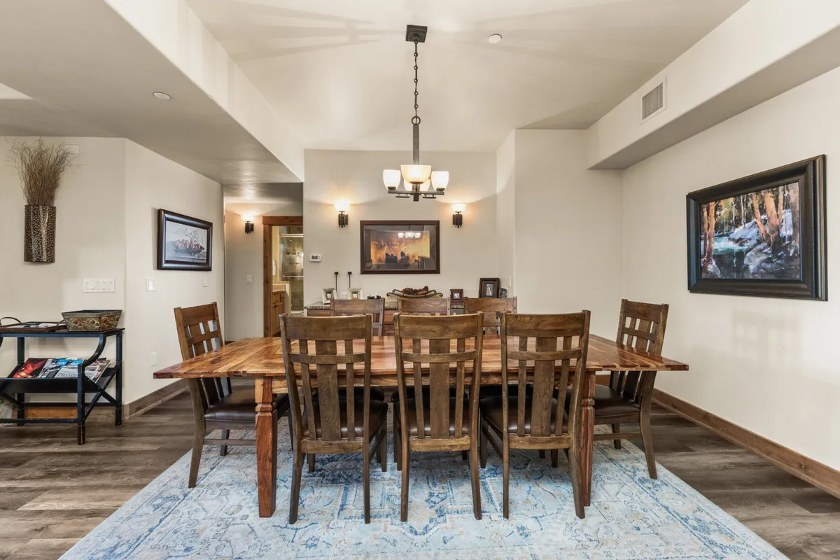 A warm dining space with a large wooden table at its center, surrounded by chairs and topped by a striking chandelier. Decorative wall art and a soft blue area rug add homeliness, while the layout is clearly designed for group meals and celebrations.