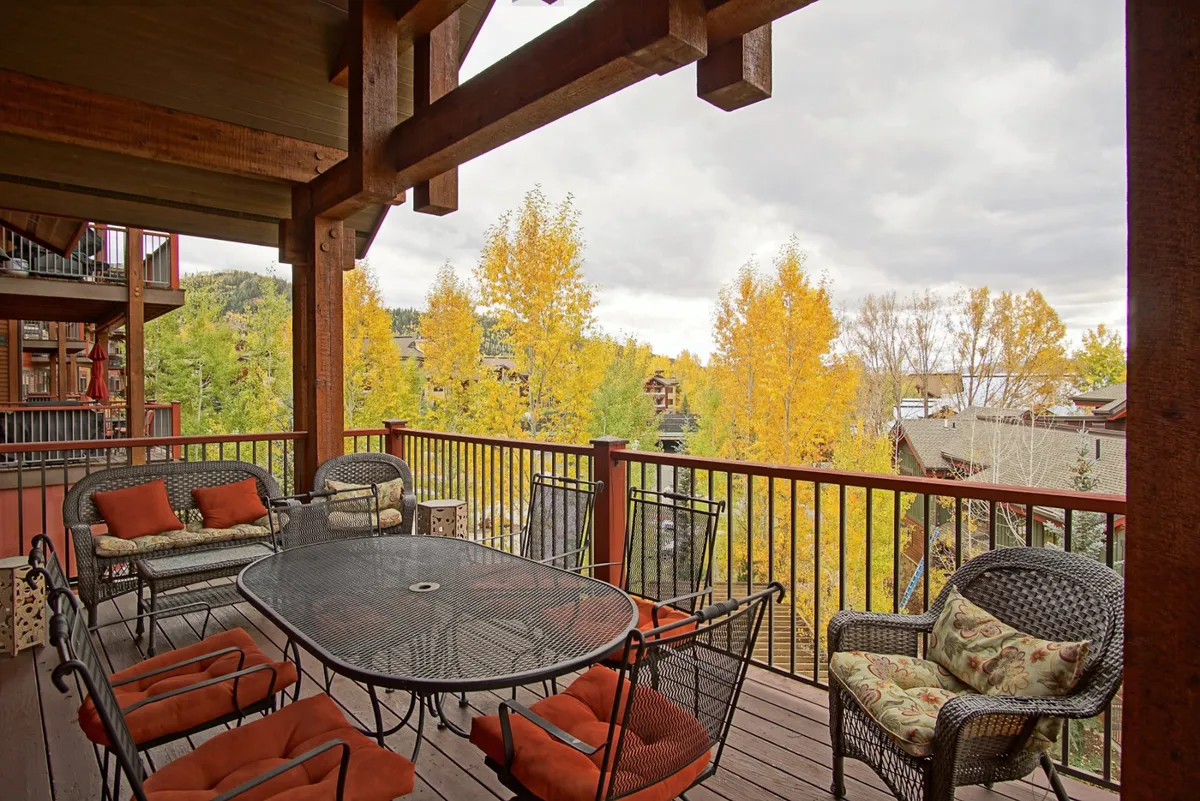 The image showcases the condo's inviting exterior deck, framed by rustic wooden beams and adorned with a metal table surrounded by colorful cushions. Vibrant autumn foliage provides a stunning mountain backdrop, inviting guests to unwind and savor the beauty of Steamboat Springs. The scene radiates a relaxed and welcoming ambiance, suggesting moments spent dining outdoors, enjoying coffee at sunrise, or gathering with loved ones as the sun sets.