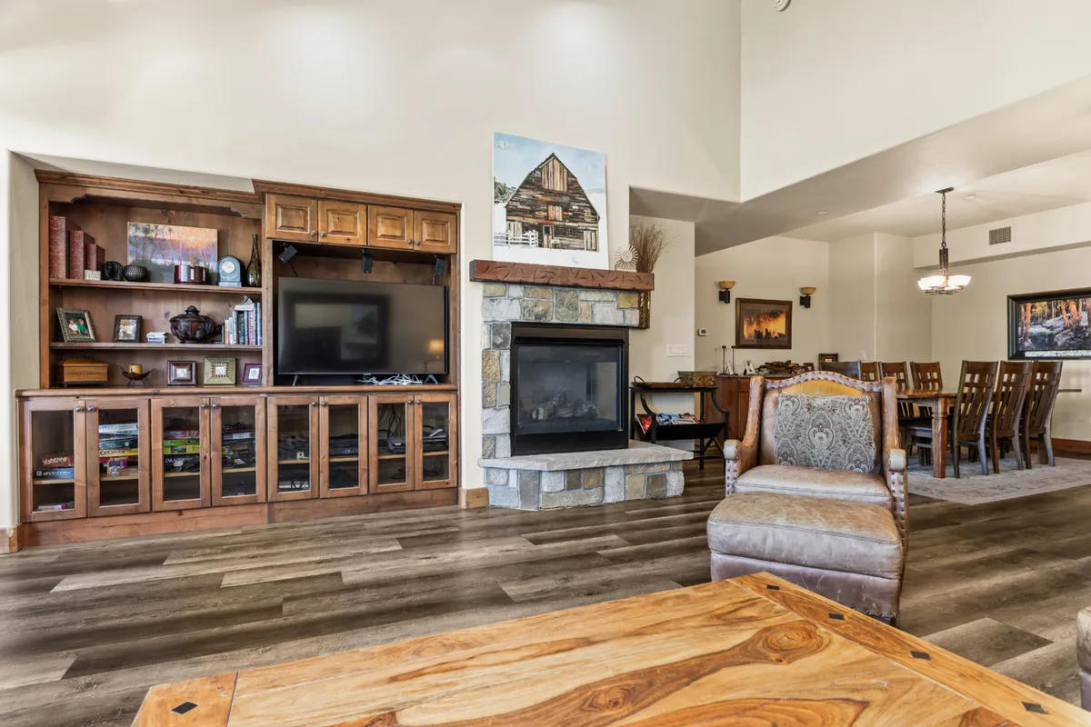 A spacious, open-concept living and dining area featuring a plush, comfortable seating arrangement centered around a striking stone fireplace. The room is accented by a wooden entertainment unit, visible dining area, and large windows that bring in abundant natural light, creating a welcoming ambiance perfect for gatherings.