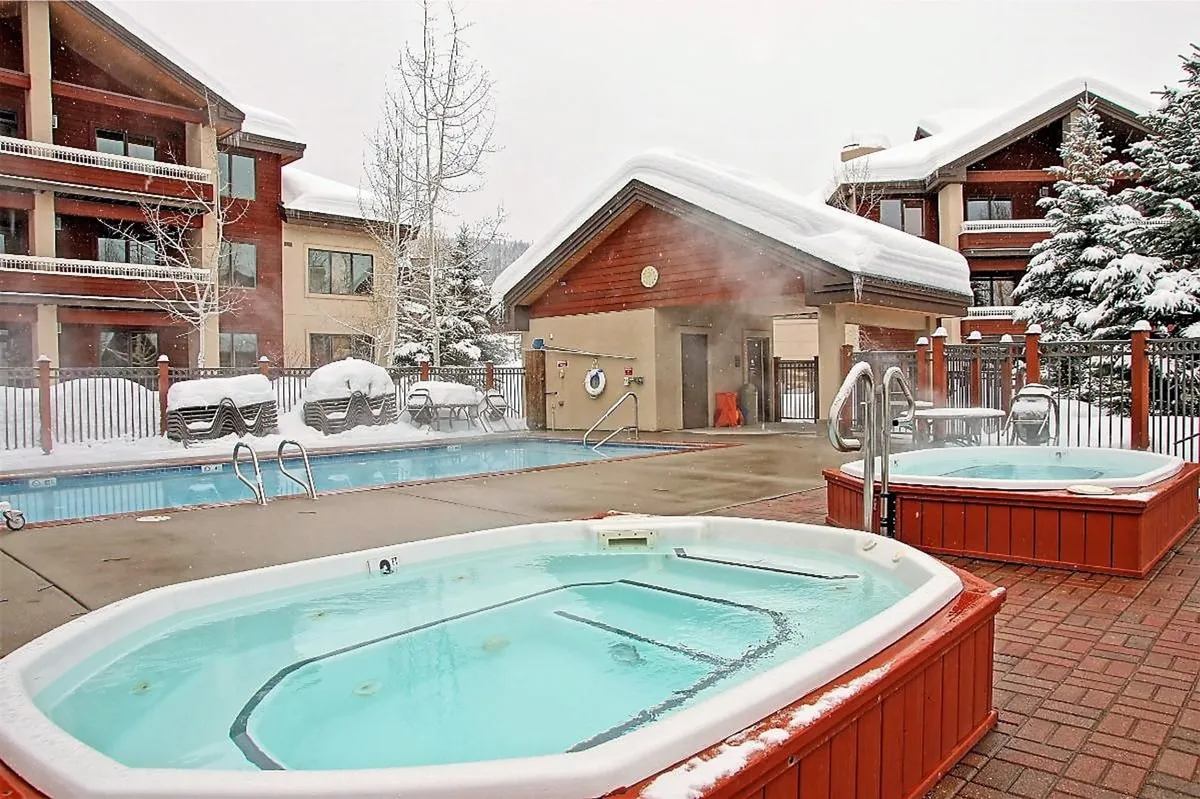 This exterior shot captures a series of inviting hot tubs and a swimming pool surrounded by snow, set against a serene backdrop. The scene exudes the unique year-round indulgence of soaking in a warm spa while taking in the mountain air, with the pool and tubs emphasizing the resort's wellness amenities.