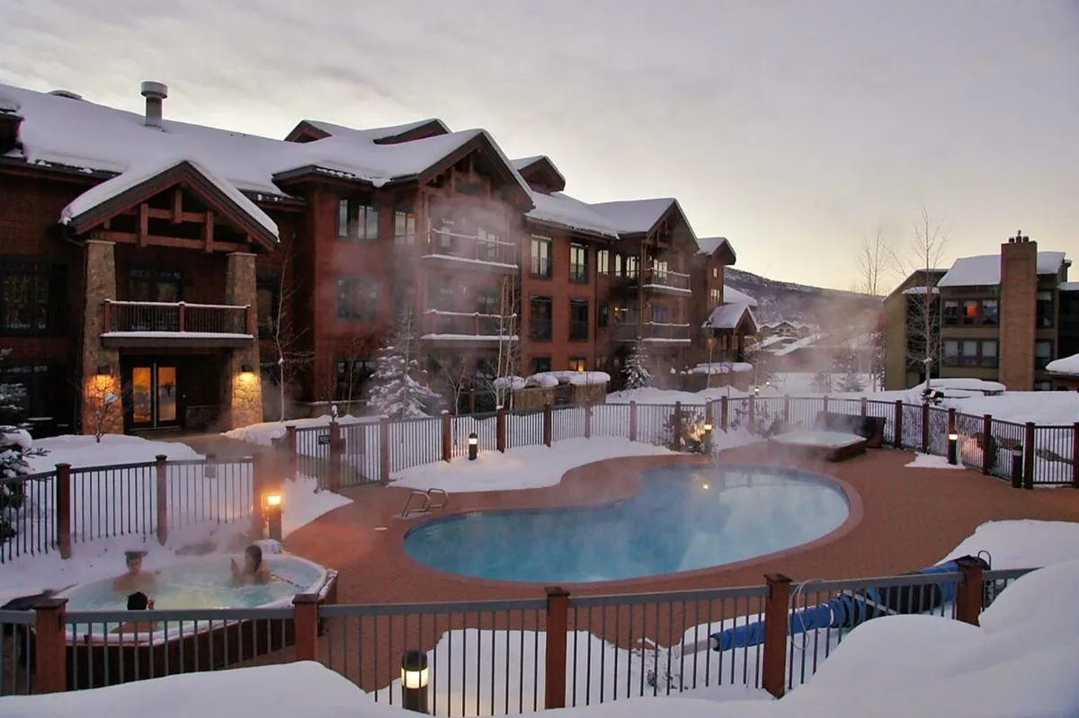 A stunning view of an outdoor heated pool and hot tubs set against a snow-covered landscape, with mountains in the distance. Guests are seen swimming, relaxing in the hot tubs, and gathering by a cozy fire pit. The scene captures the resort’s vibrant winter ambiance and highlights the year-round utility of the amenities, with steam rising from the water and lights creating a warm glow.