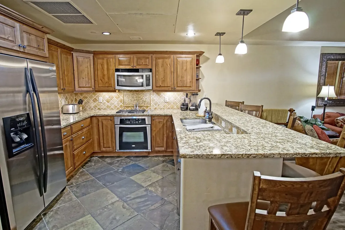 A warmly lit kitchen featuring a spacious granite countertop, wood cabinetry, and stainless steel appliances. Bar stools are pulled up to the counter, inviting casual conversations or snacks while cooking. The kitchen exudes both style and function, with ample prep space and a welcoming atmosphere for gatherings.