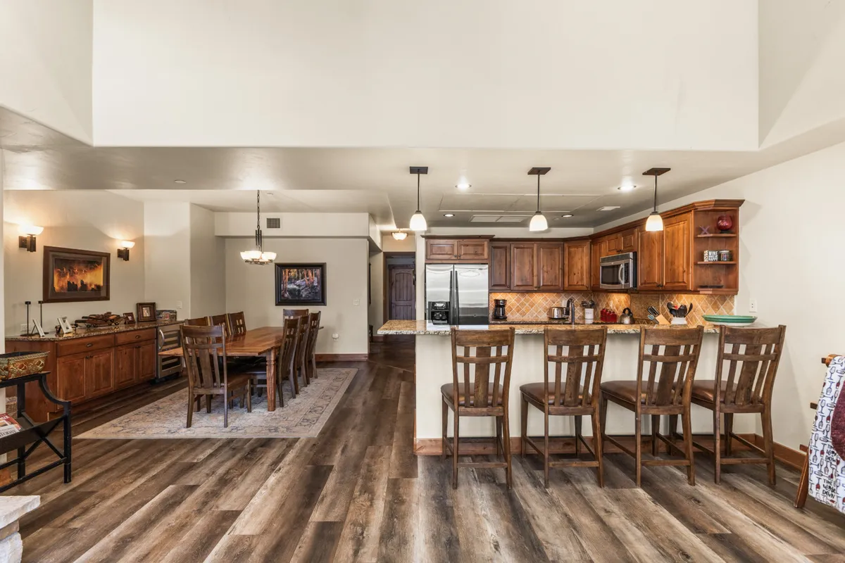 A broad view of the open kitchen and adjacent dining area. The kitchen island is paired with bar stools, and the large wooden dining table is set for a group. Wooden cabinetry and tasteful artwork create a cozy and stylish ambiance, ideal for both formal meals and casual brunches.